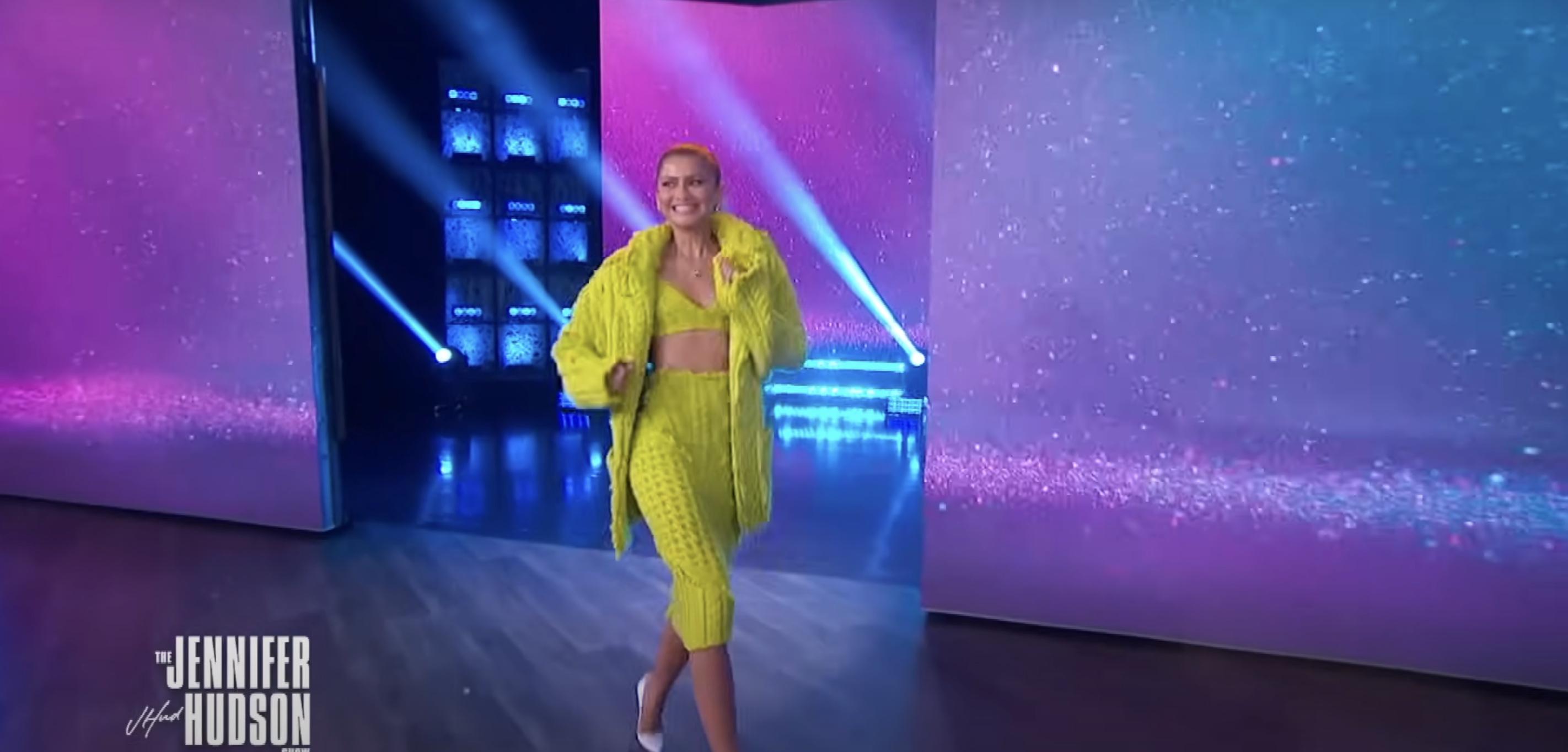 Person in a textured yellow outfit with a crop top and pants, standing on a stage with lit-up background