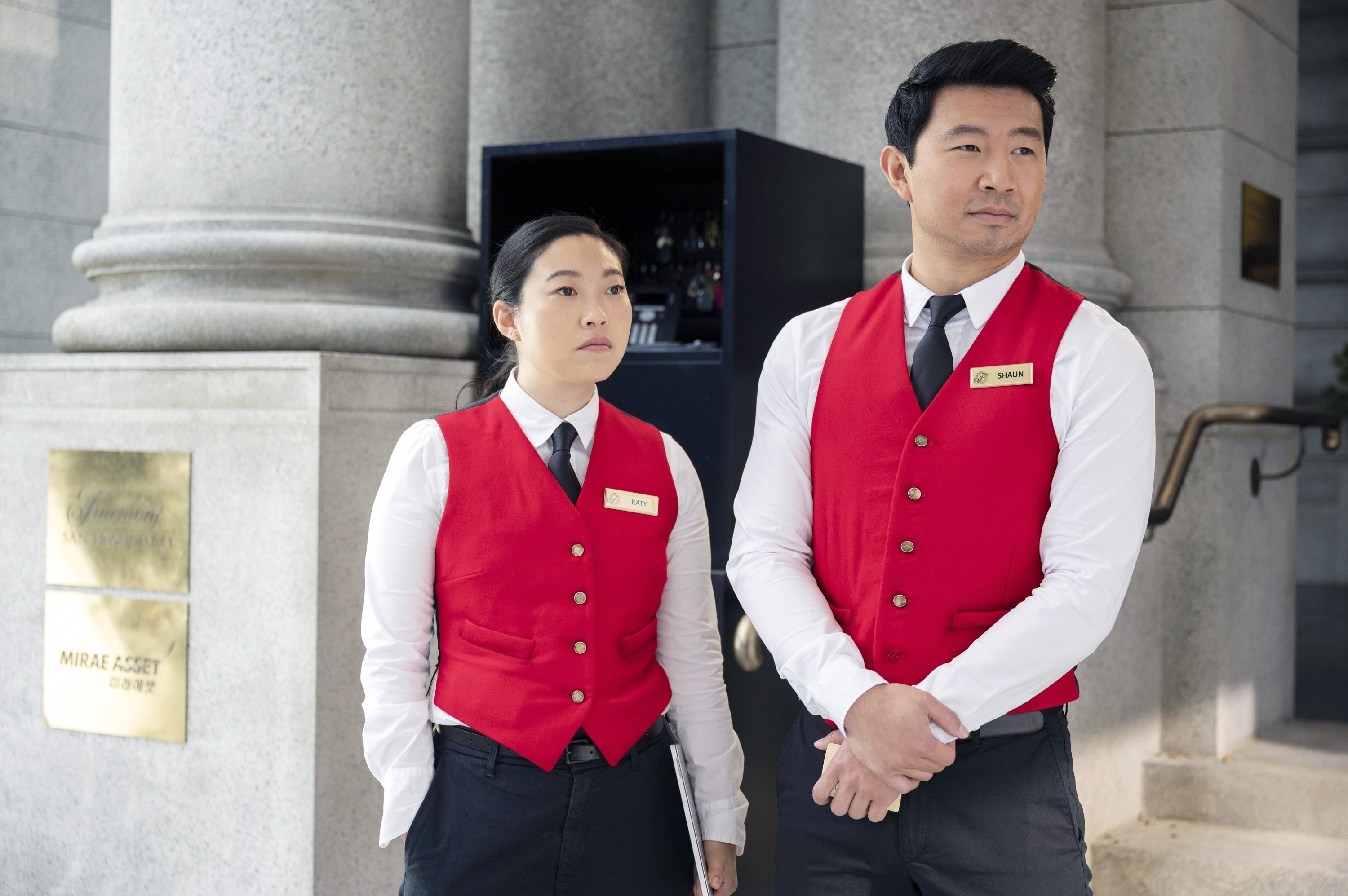 in a scene, Awkwafina and Simu portraying valet attendants in vests and name tags stand near a building