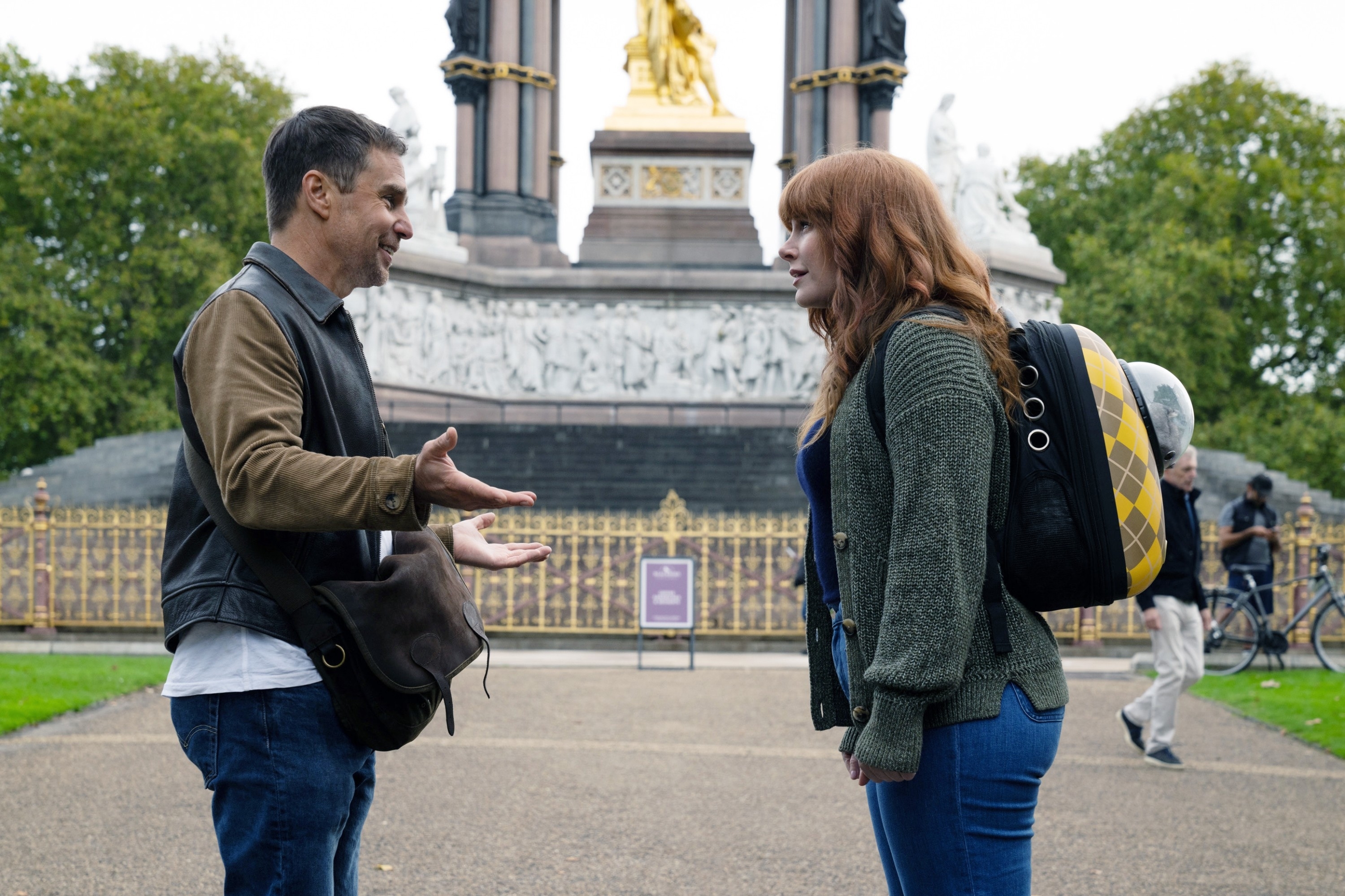 Two actors in a scene, Sam gesturing while talking to a smiling Bryce with a backpack, outdoors near a statue