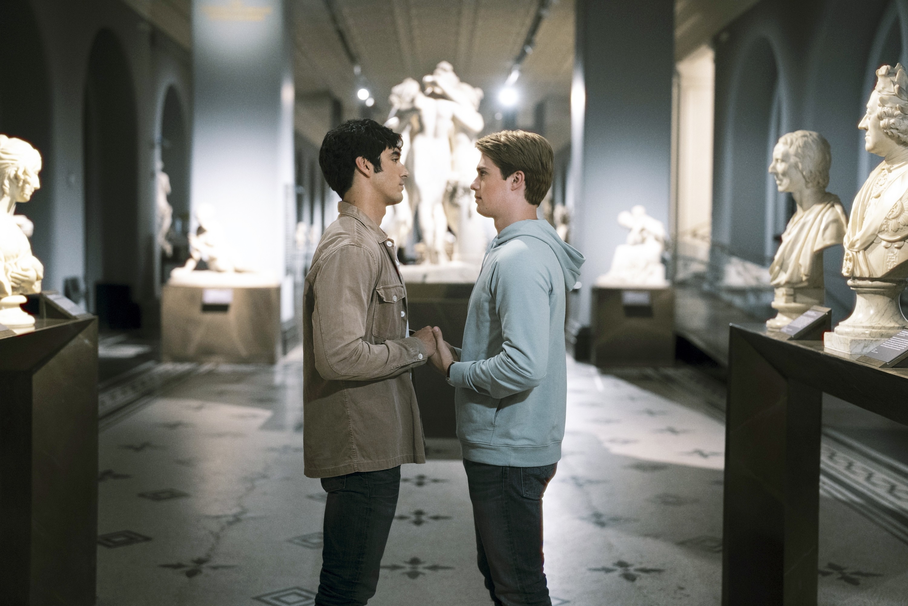 in a scene, Nicholas and Taylor portraying a couple holding hands in a museum, facing each other with classical statues in the background