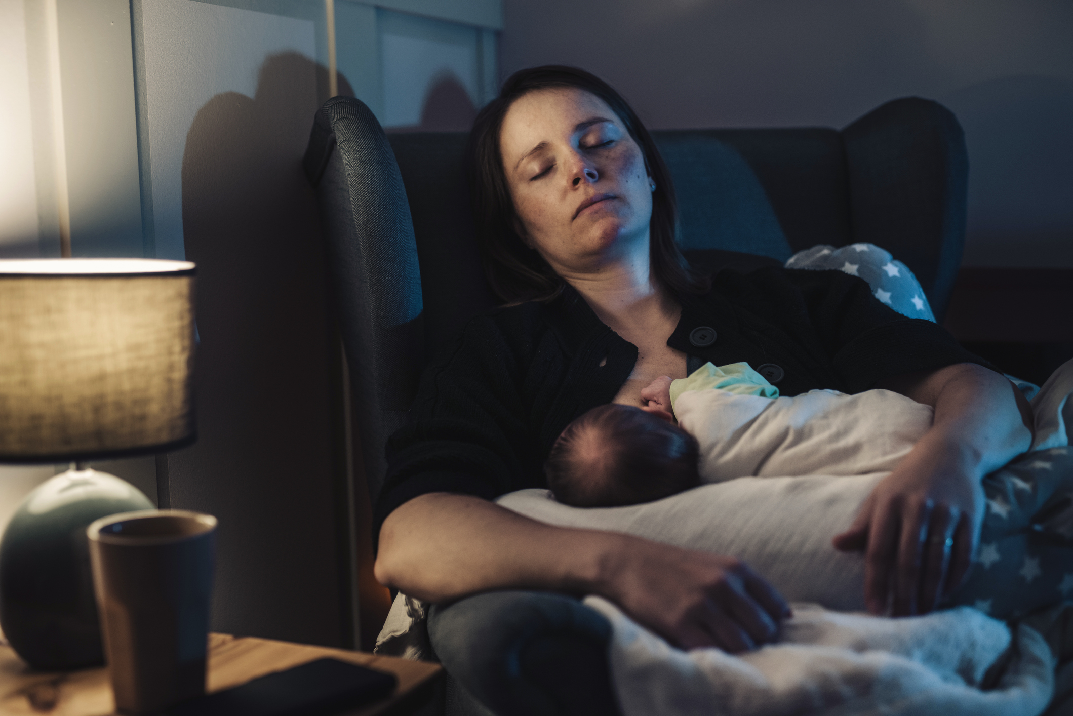 A tired mother with her sleeping baby resting on her chest in a dimly lit room, conveying a moment of parental care
