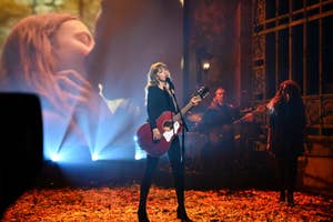 Taylor Swift performing onstage with a guitar, with band members and autumn leaves on the floor
