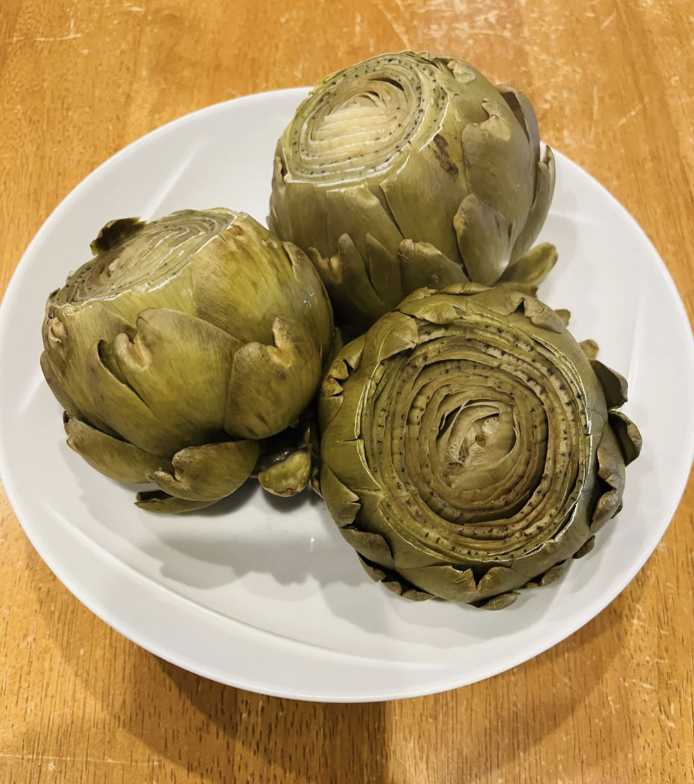 Three whole artichokes on a white plate atop a wooden surface