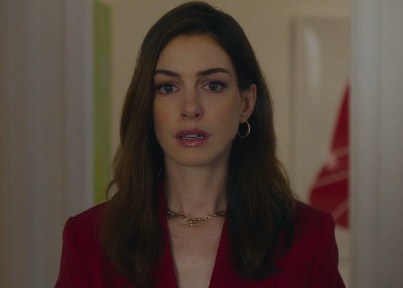 Anne Hathaway con traje rojo y collar, expresión seria, en interior