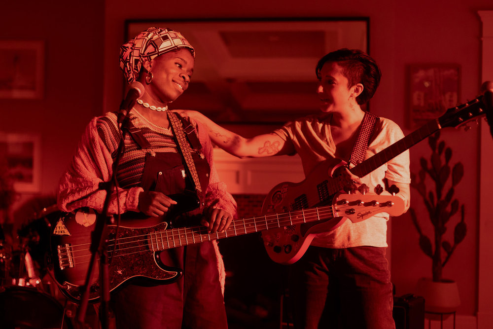Two musicians, one with a headscarf and overalls, and the other with a short haircut, holding a guitar, engage warmly onstage