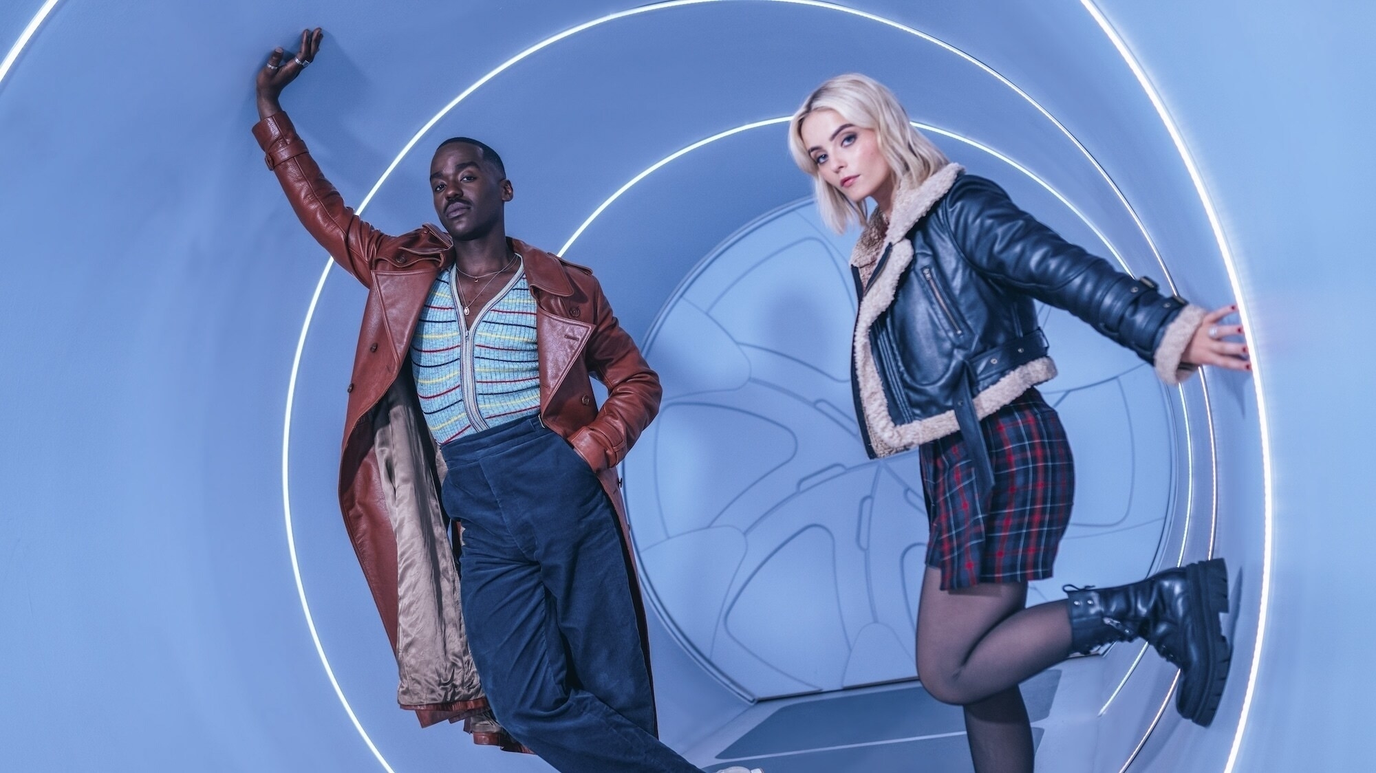 Two actors pose in front of a futuristic backdrop, man in a brown jacket and woman in a black leather jacket and plaid skirt