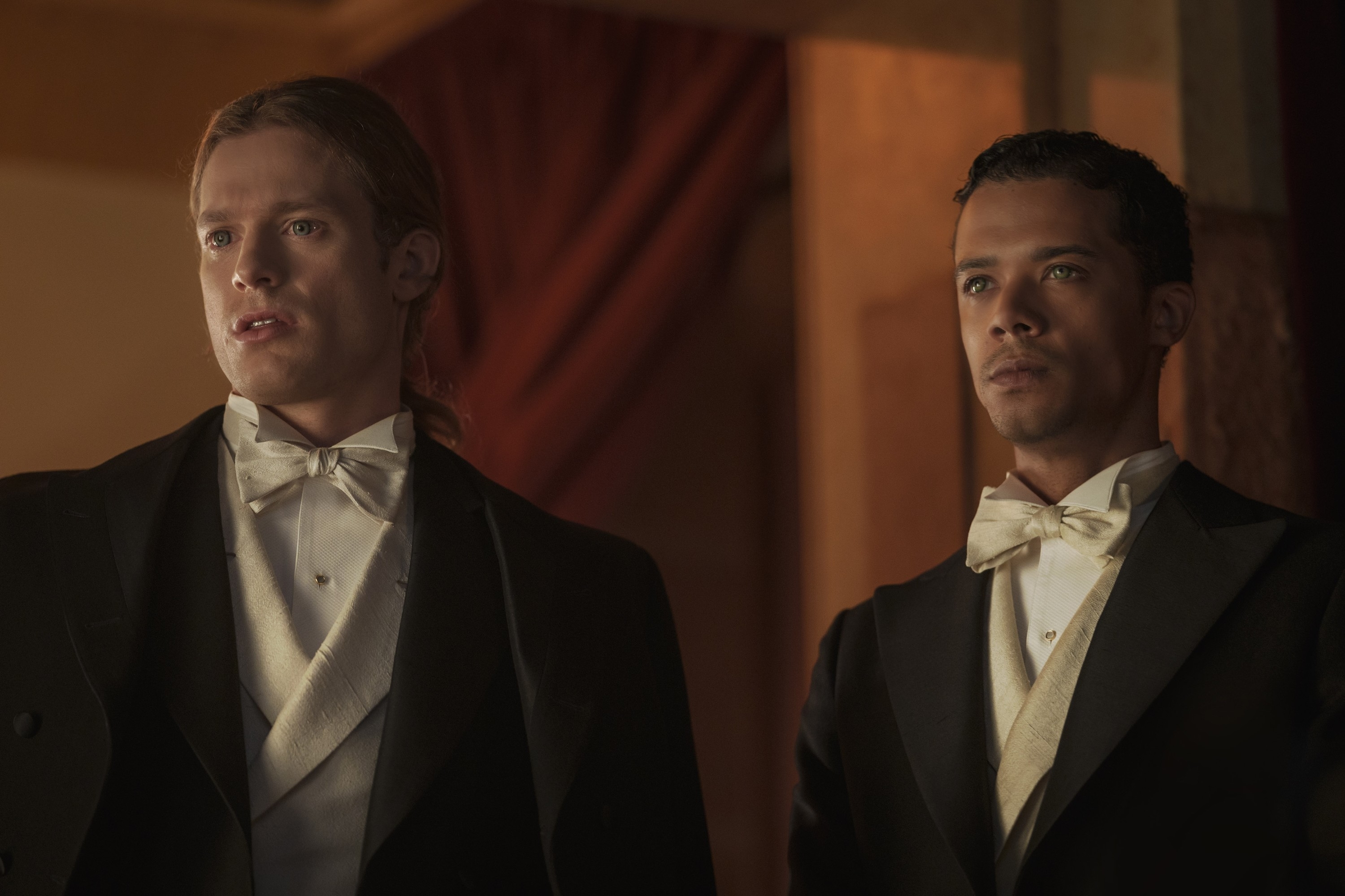 Two men in formal black tuxedos with bow ties, looking intently to the side, in a dimly lit setting