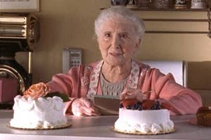 Elderly woman as Annie Camden in a kitchen with two cakes, looking pleasantly surprised