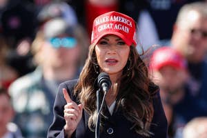 Woman in "Make America Great Again" hat speaks at an event, gesturing with her index finger raised