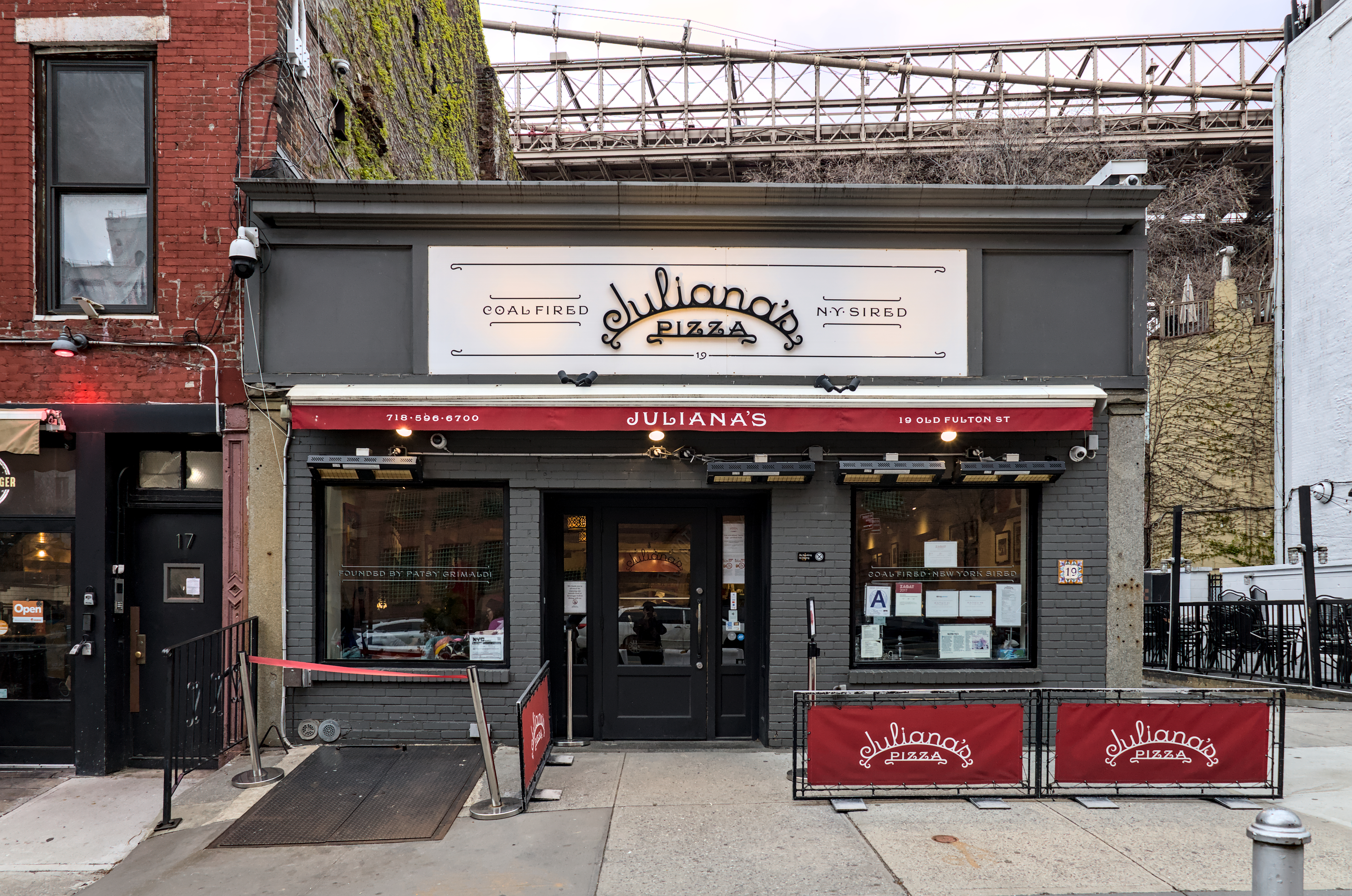 Exterior view of Juliana&#x27;s Pizza under a bridge, with signage, outdoor seating, and dual entrance doors
