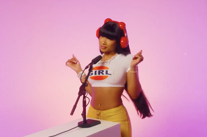 Person on pink backdrop by mic wearing a cropped top with 'GIRL' print, yellow pants, and red hair accessories