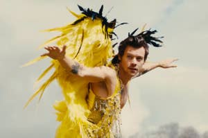 Harry Styles in a yellow feathered outfit with beaded details, arms outstretched, against a sky backdrop