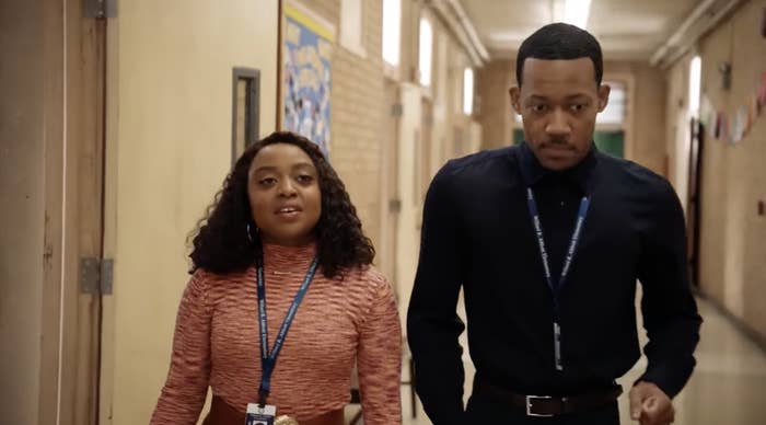 Janine and Gregory from Abbott Elementary walking down a school hallway; a woman in a ribbed top and a man in a shirt and tie
