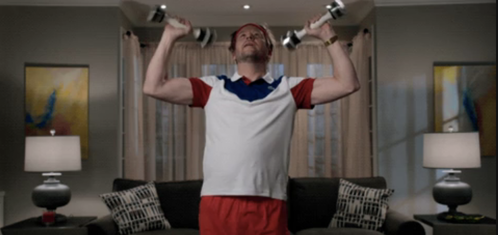 Man in casual workout gear lifts dumbbells indoors; focused expression as he exercises