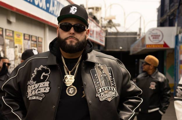 Man with beard wearing a black jacket with embroidered patches, a cap, and gold chain