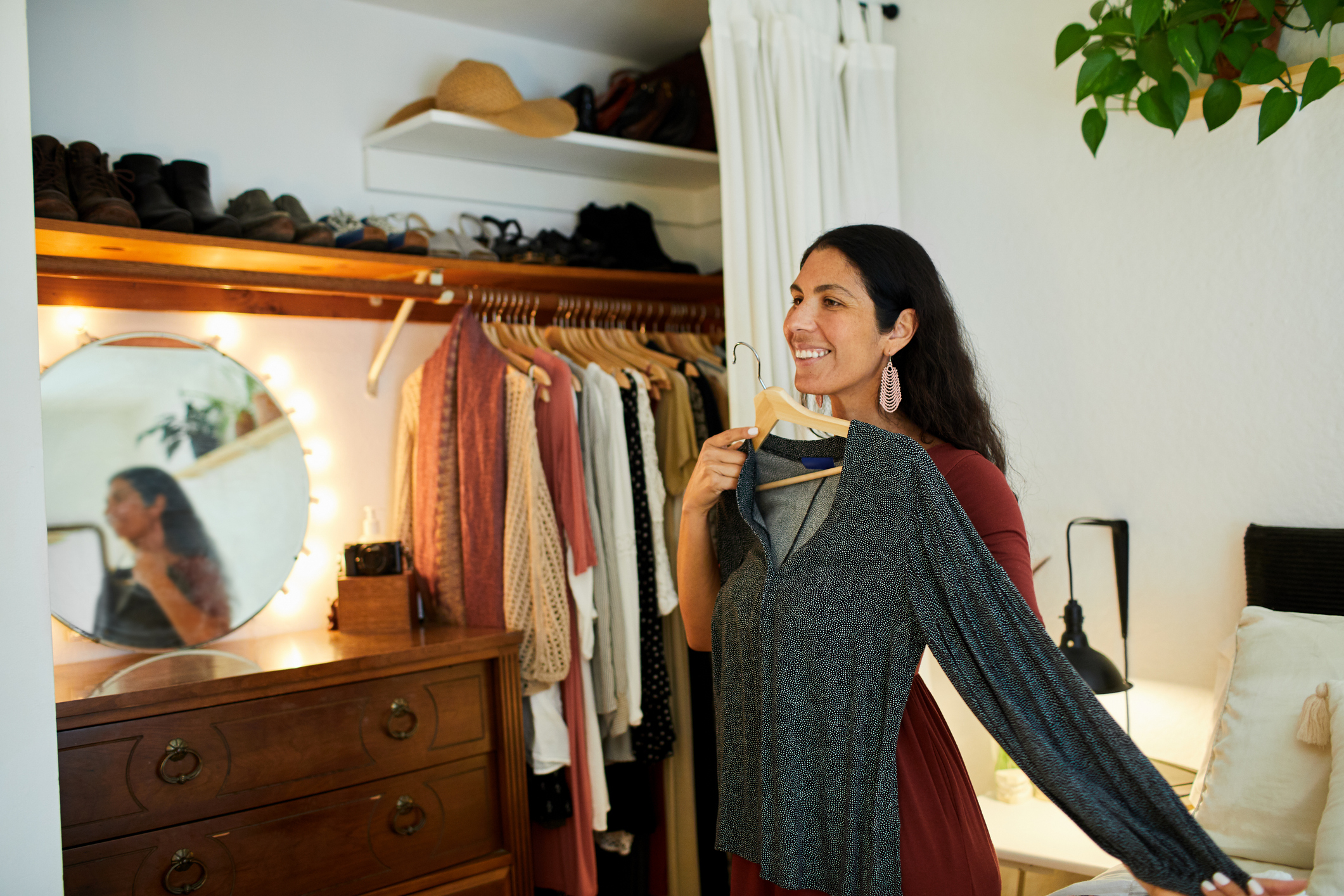 Woman smiling, holding a shirt, standing by a mirror and wardrobe, reflecting environmentally friendly fashion choices