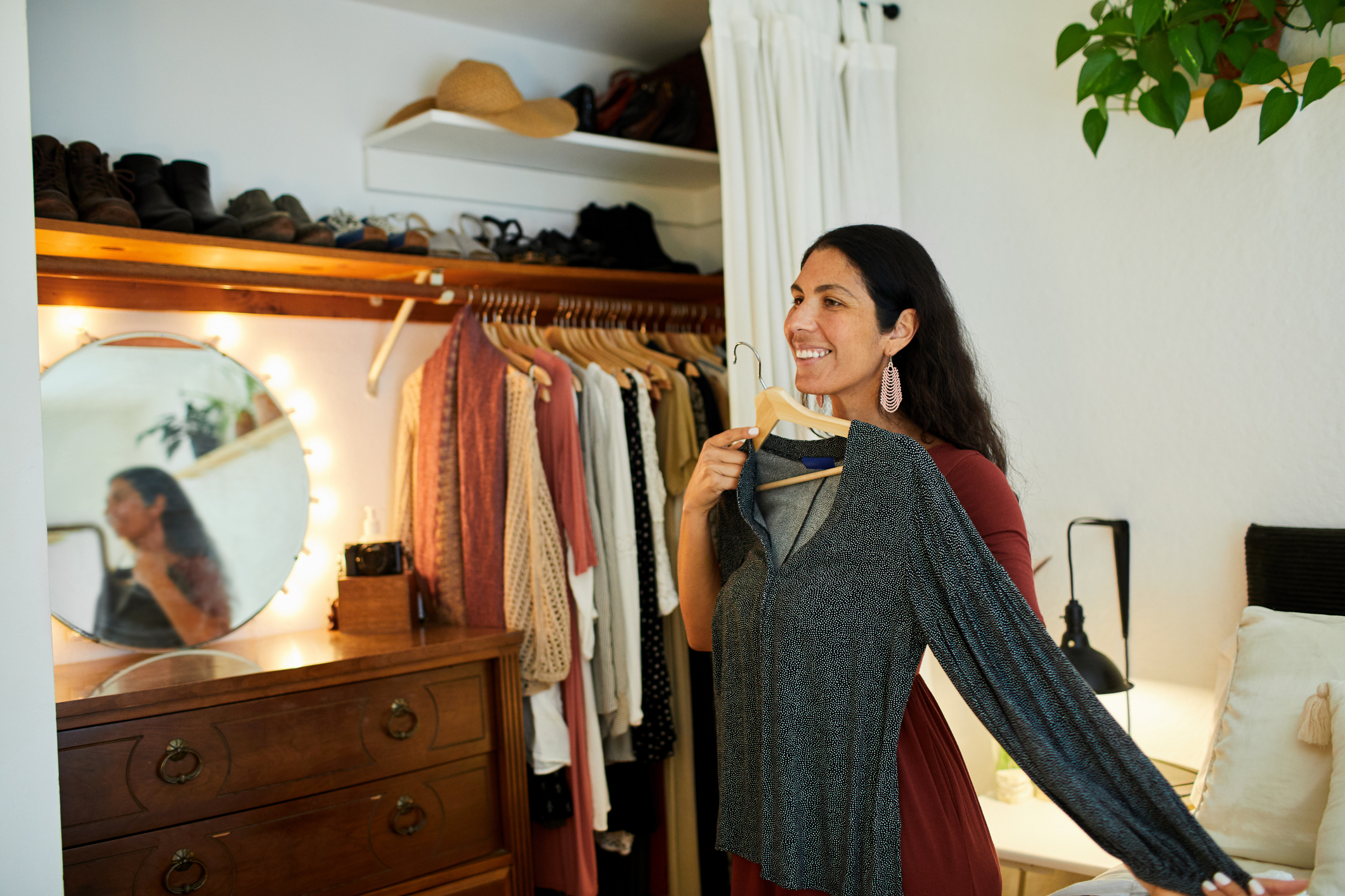 Woman smiling, holding a shirt, standing by a mirror and wardrobe, reflecting environmentally friendly fashion choices