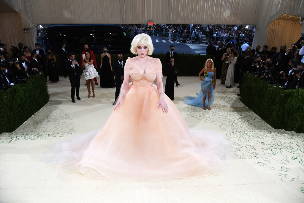 A person in an extravagant gown with a voluminous skirt and bow detailing stands on a staircase at an event. Background figures include photographers
