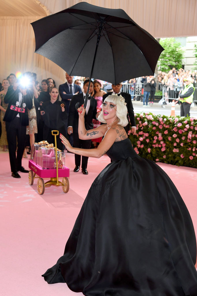 Lady Gaga on the red carpet holding an umbrella, with a long, elegant gown and a wagon with props behind her