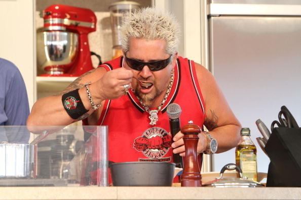 Guy Fieri tasting food while cooking, wearing a red tank top, with sunglasses and jewelry
