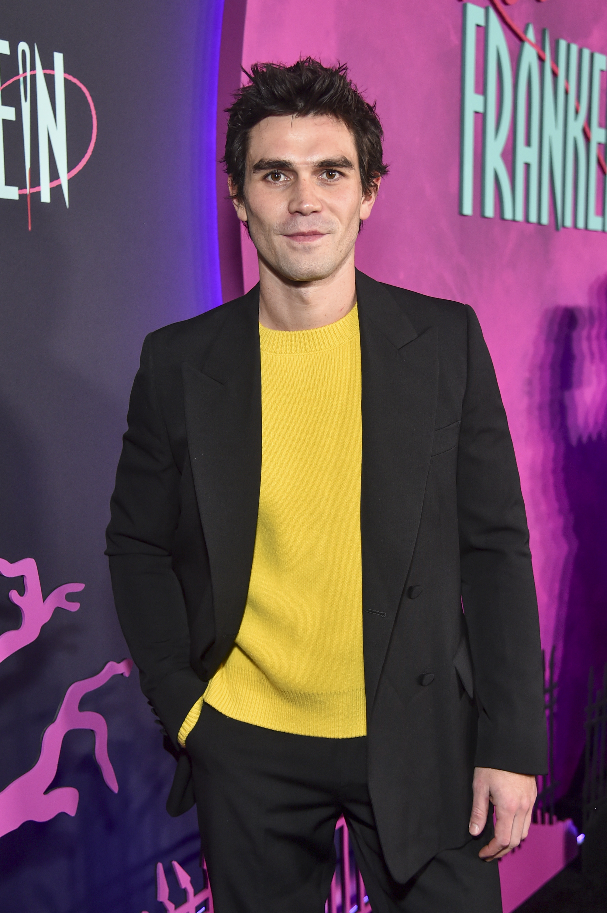 KJ Apa in a black blazer over yellow sweater poses against promotional backdrop
