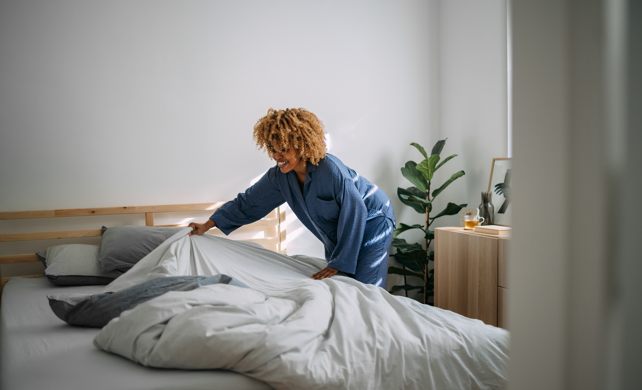 Person making bed in a bright room, dressed in a casual robe with a relaxed expression