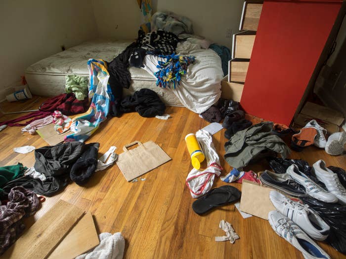 A messy bedroom with clothes and shoes scattered on the floor, unmade bed, and personal items around