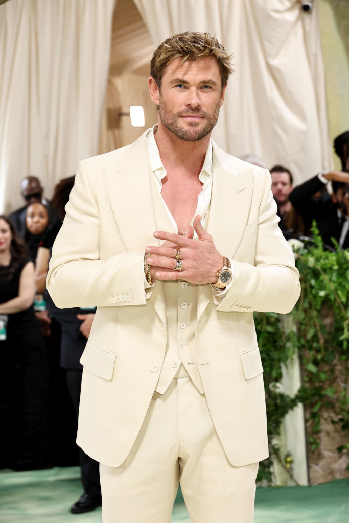 Chris Hemsworth in a cream suit and open shirt at an event, looking at the camera