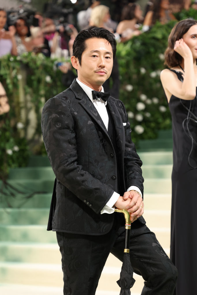 Steven Yeun stands on the red carpet in a detailed tuxedo with a pattern, holding a cane