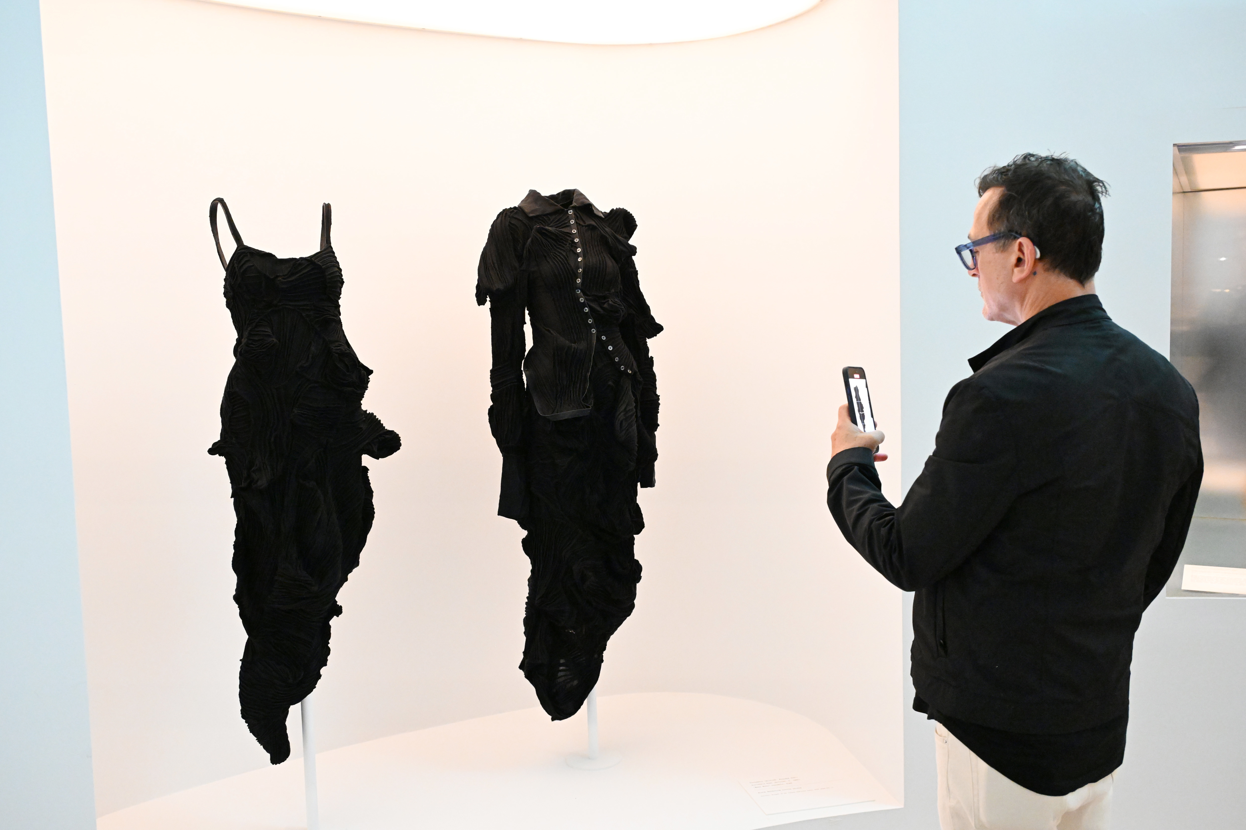 Man views two textured dresses on display at an exhibition, one with a ruffled design, capturing them with his smartphone