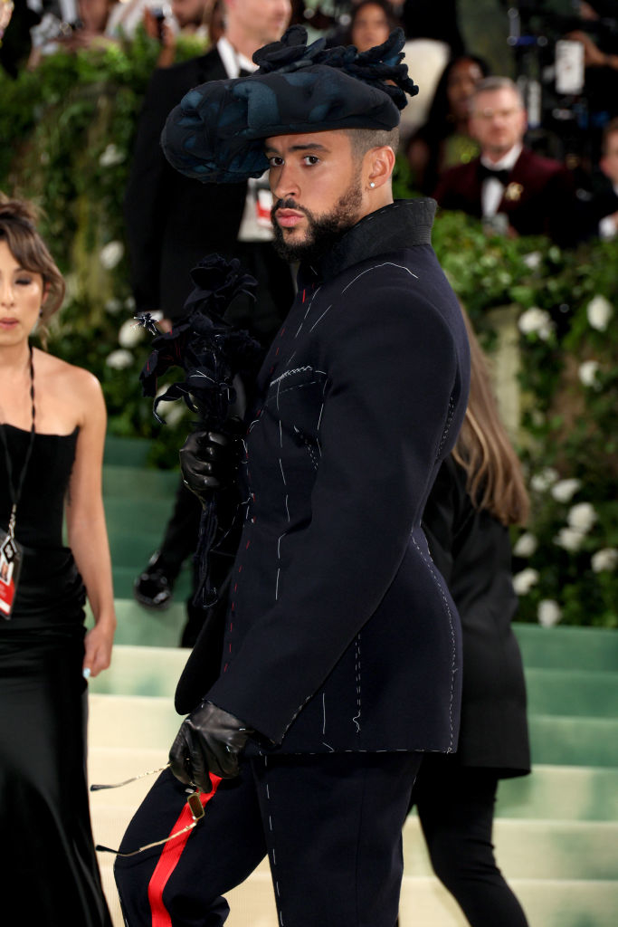 Bad Bunny in a black outfit with a prominent hat and gloves on a red carpet