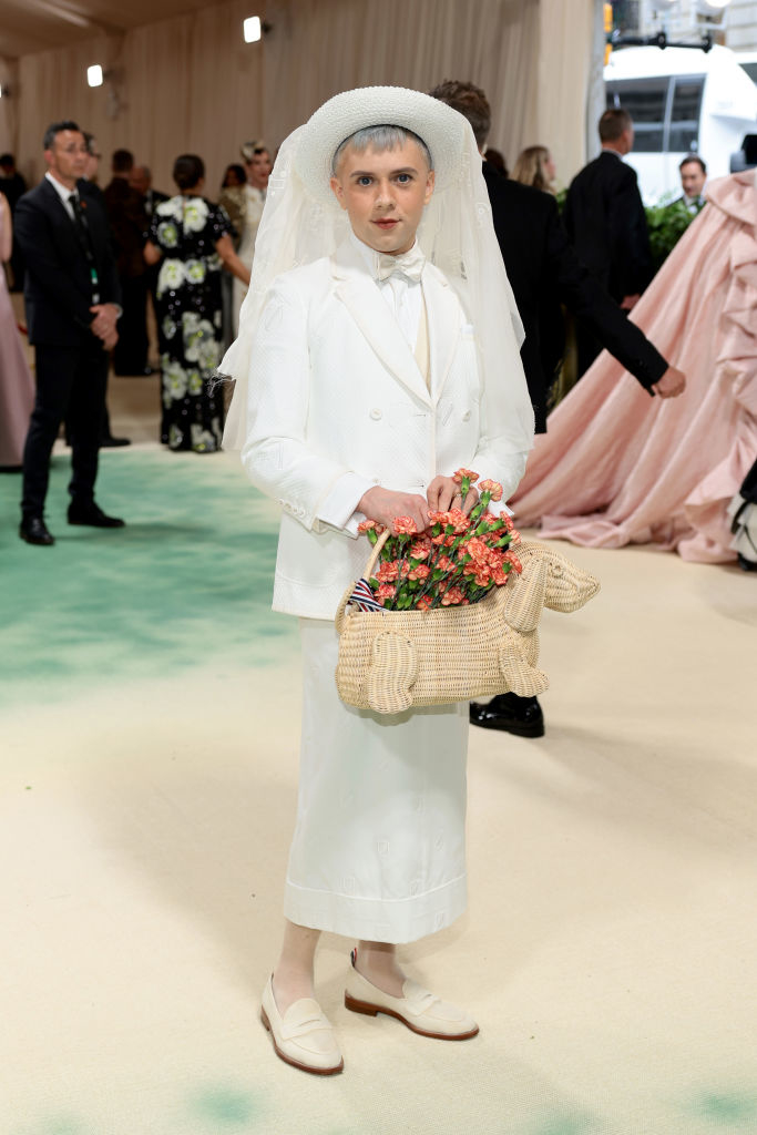 Cole Escola in a white suit with matching hat and veil holding a basket of flowers at an event