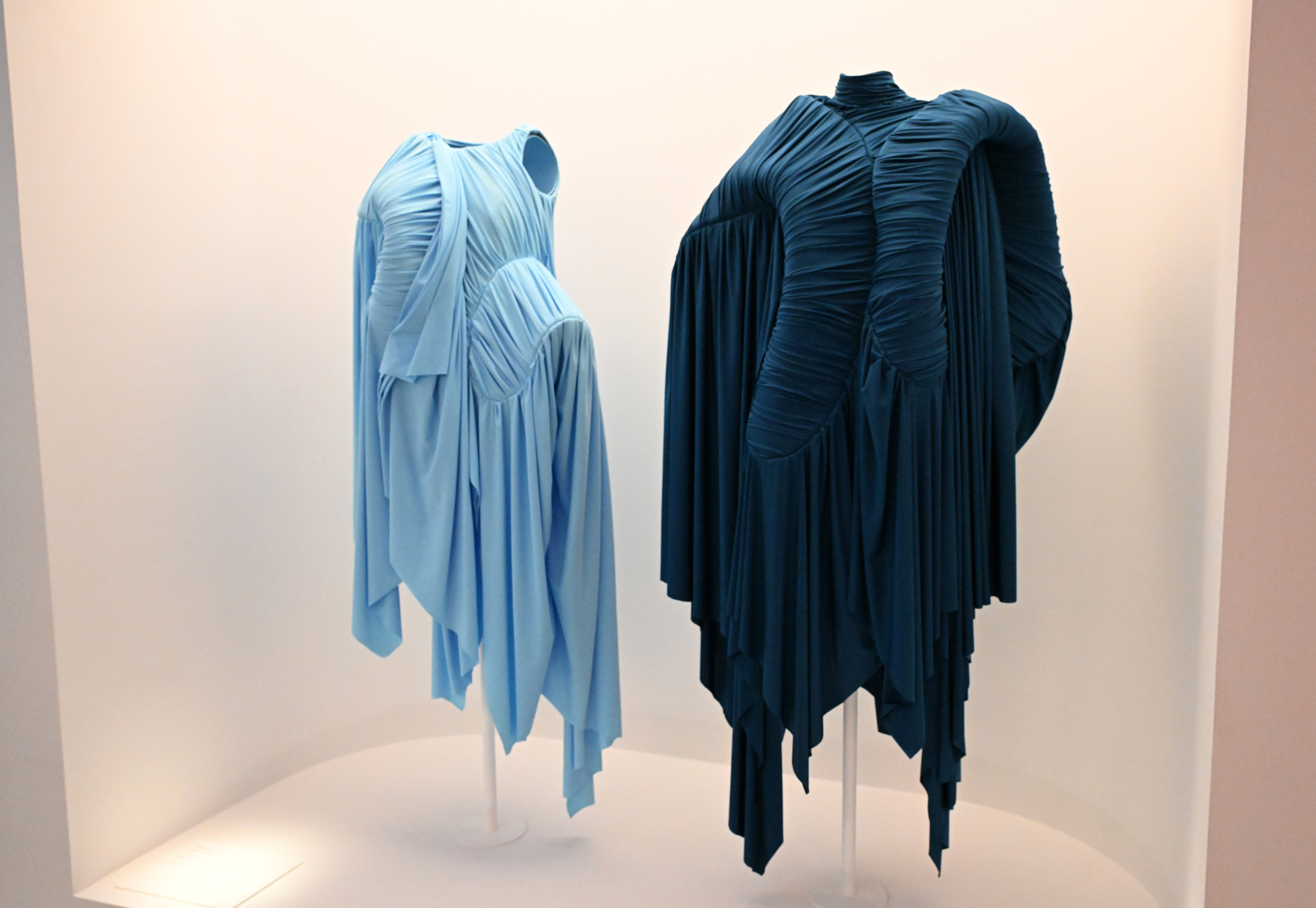 Two pleated dresses on display, one light blue and one dark blue, with unique asymmetrical designs