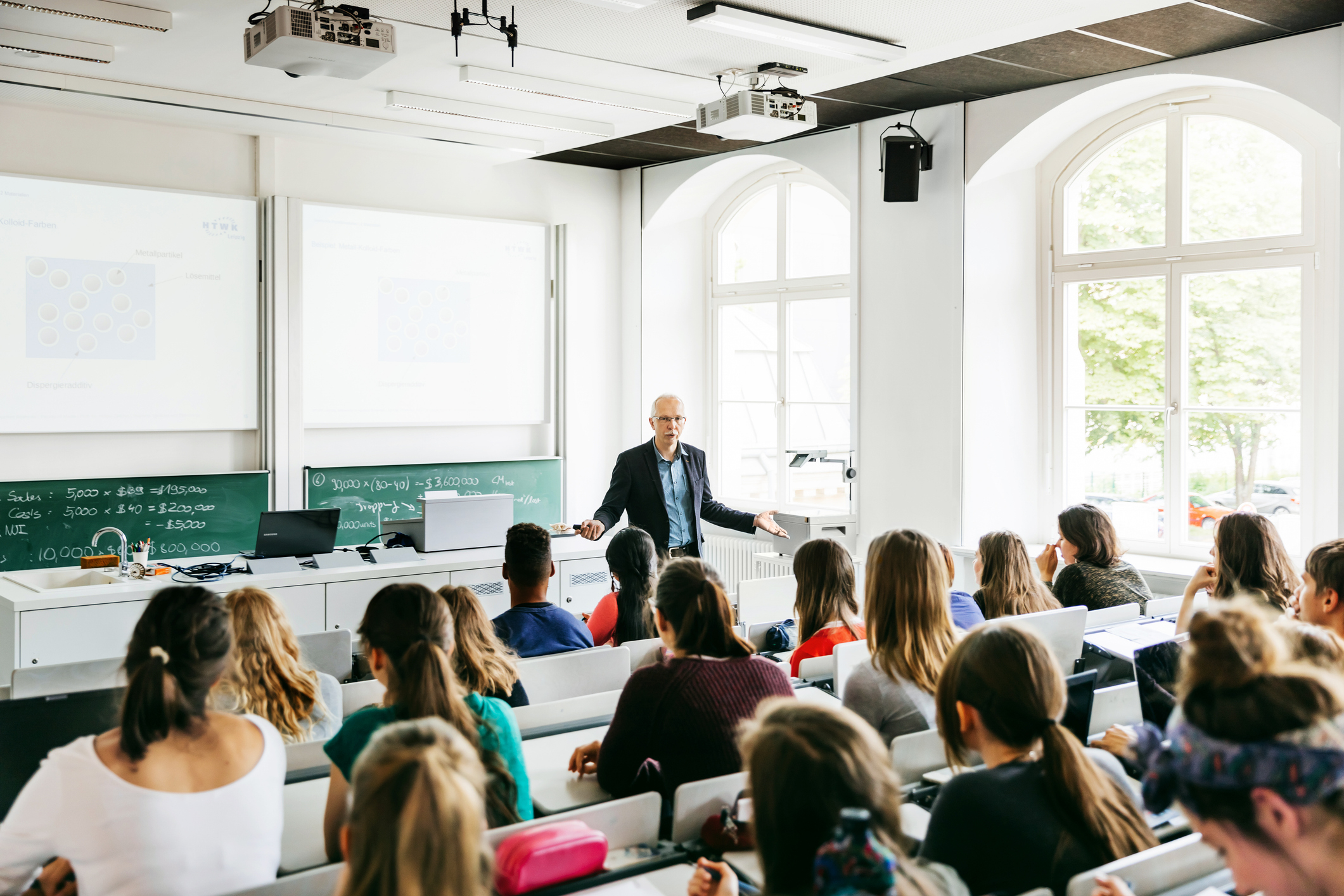 Professor lecturing in a university classroom filled with students, with projection screens visible