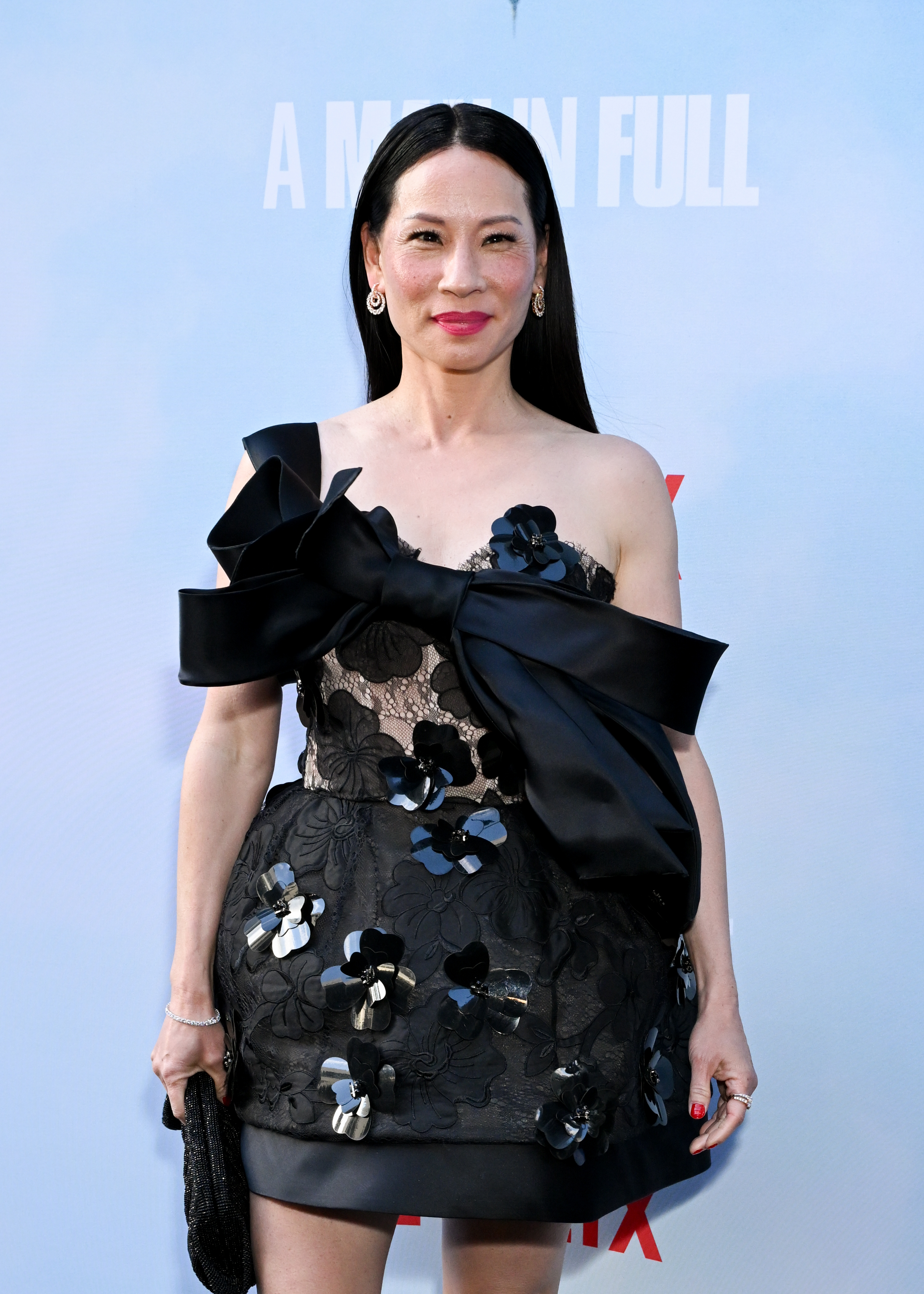 Lucy Liu in a black lace dress with large bow and floral details poses on event backdrop