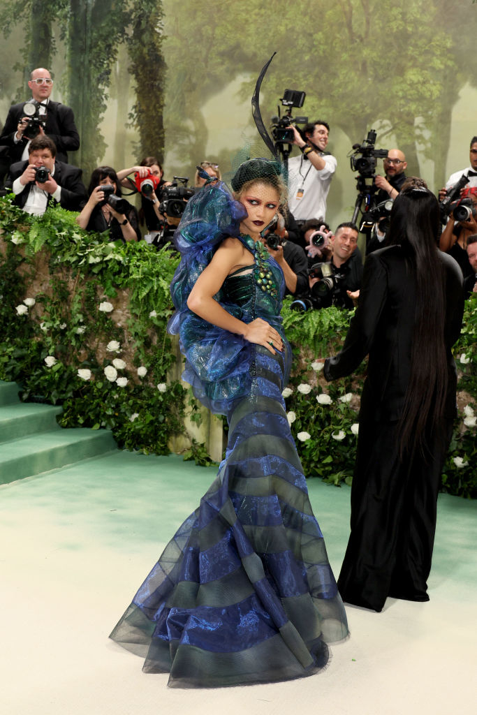 Zendaya in avant-garde  dress with ruffled layers and horn-like headpiece poses on event carpet, photographers in background