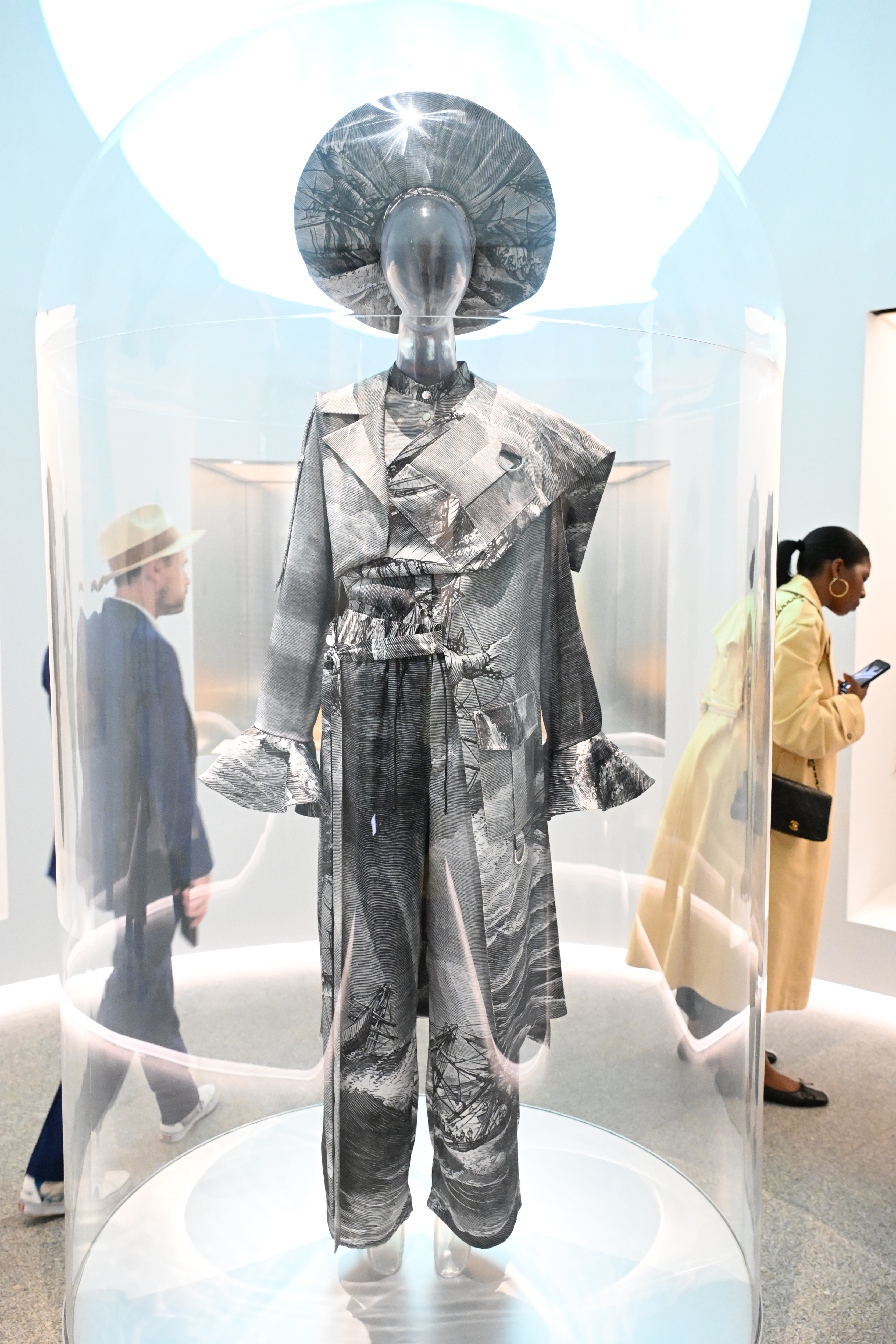 Mannequin displaying a patterned suit with a hat, exhibited in a glass case, with people observing in the background