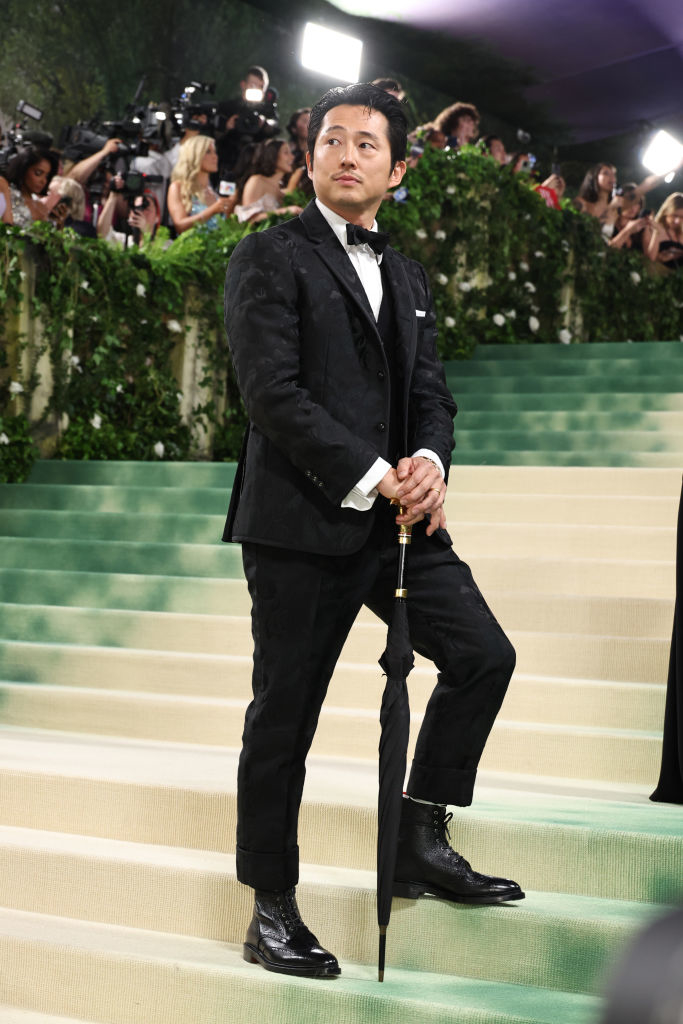 Steven Yeun in a black tuxedo with bow tie holding a cane, standing on steps with onlookers in background