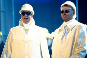 Two men in white outfits and sunglasses stand side by side onstage