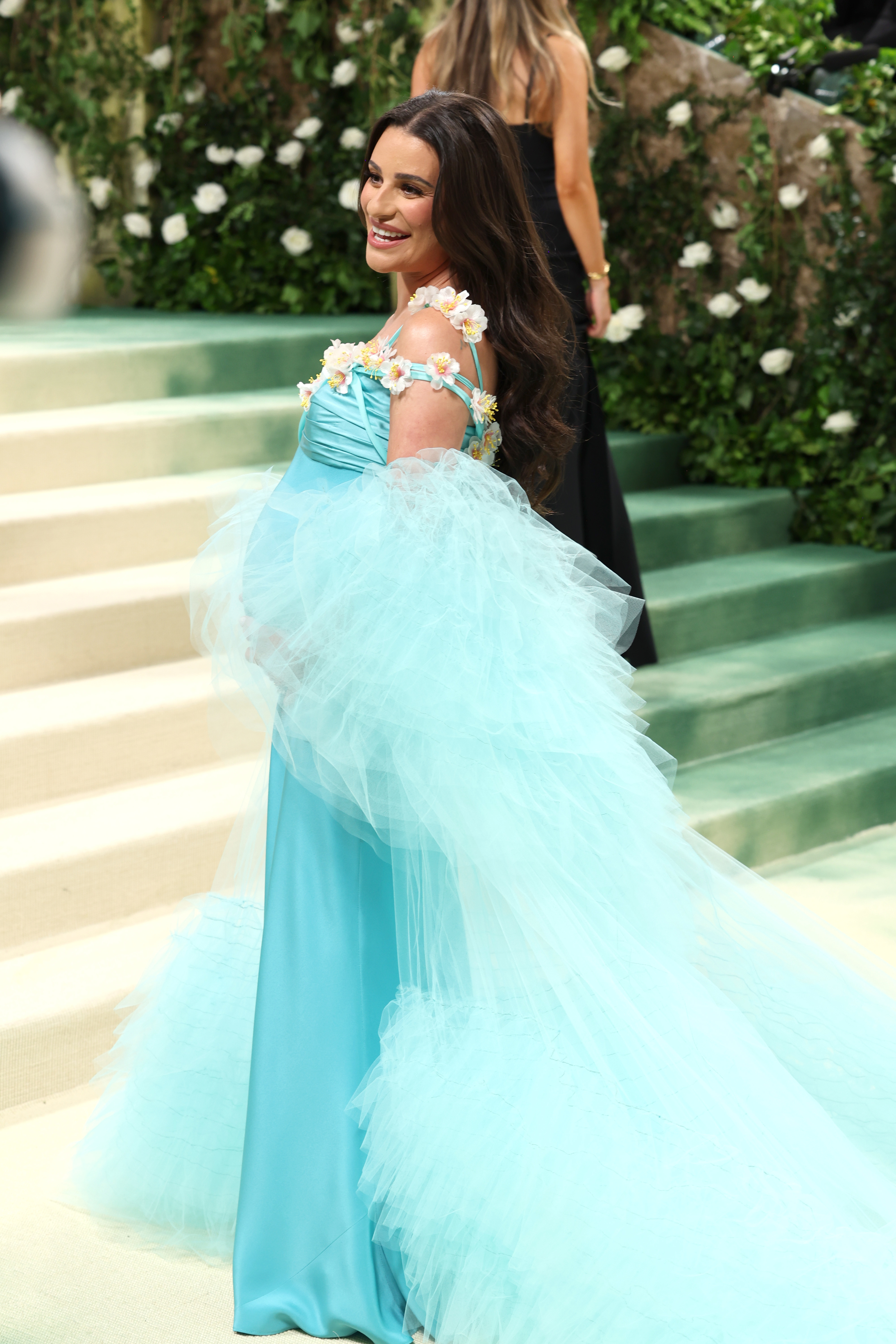 Lea Michele en un evento, sonriente, con un vestido largo con adornos florales y una capa de tul