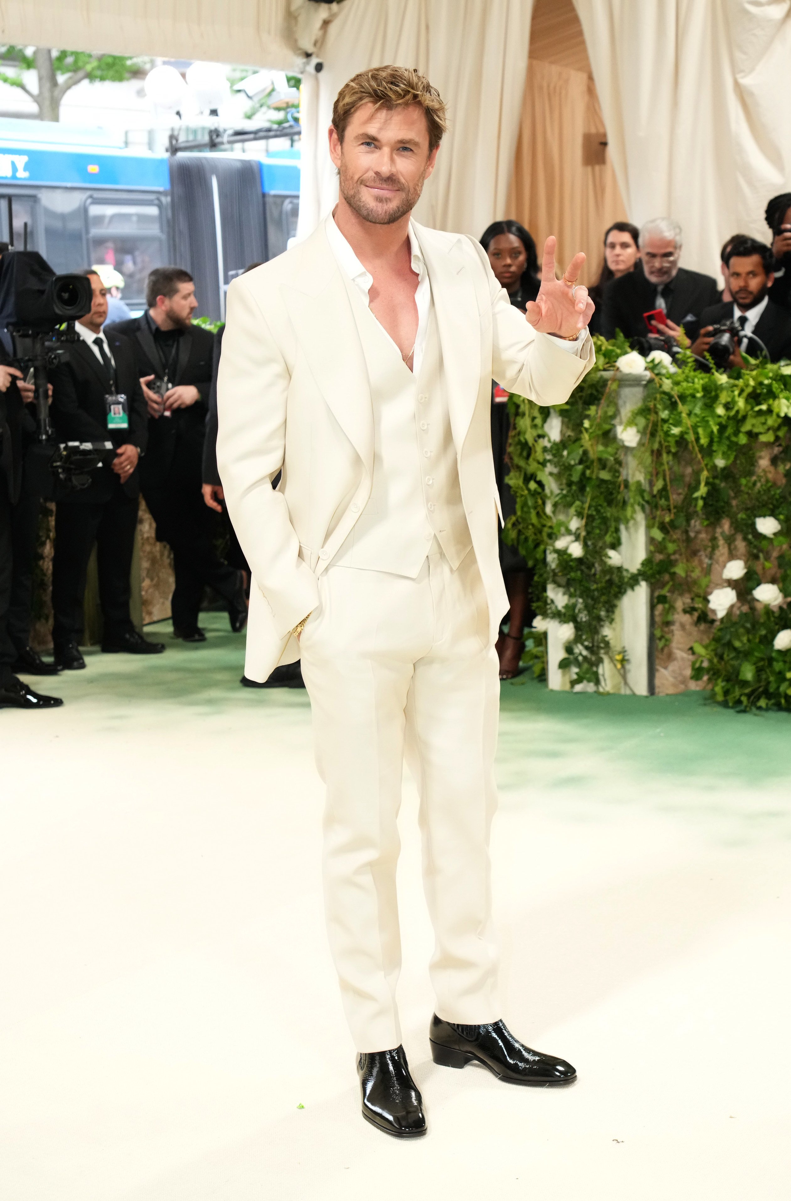 Chris Hemsworth in a white suit and black shoes flashing a peace sign at an event
