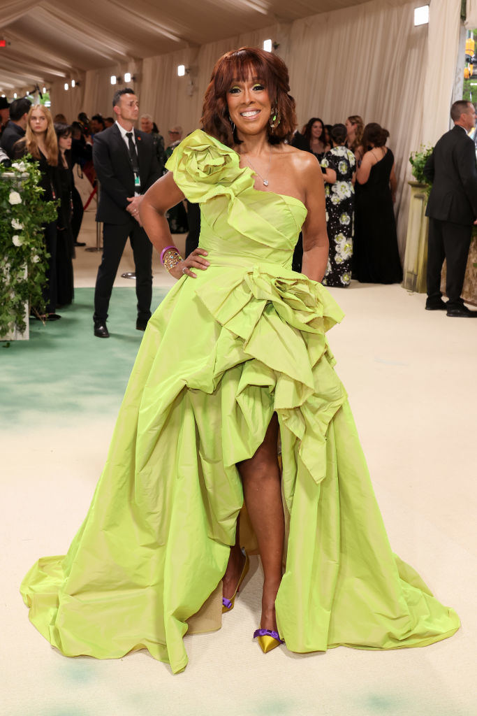 Gayle King in a lime green high-low gown with ruffles, purple heels, smiling on a crowded event floor