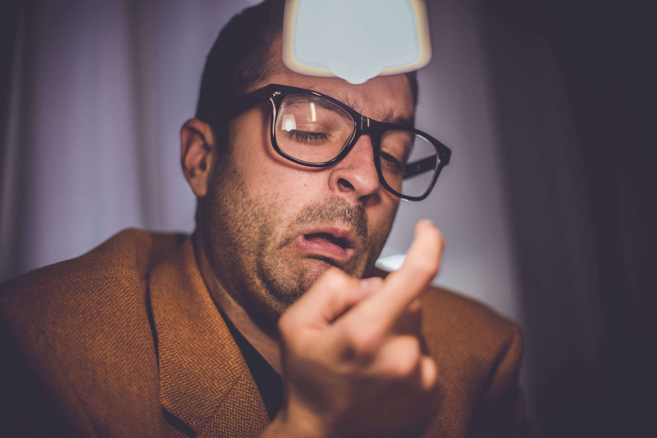 Man with glasses looking at his bandaged finger, wearing a brown jacket, surprised expression