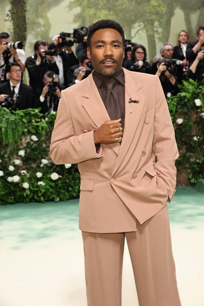 Donald Glover in elegant suit poses with hand on lapel at an event with photographers in background