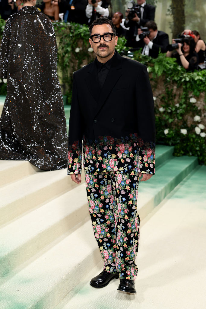 Dan Levy in black jacket and floral pants on stairs at an event