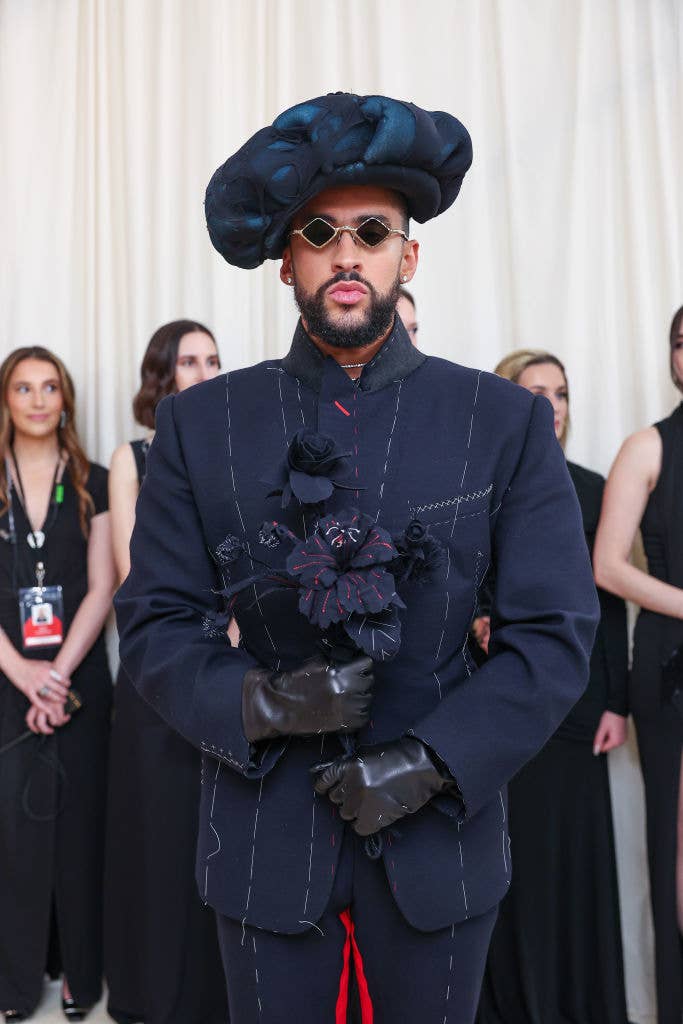 Person wearing a large hat, dark suit with flower detail, and gloves; others in background