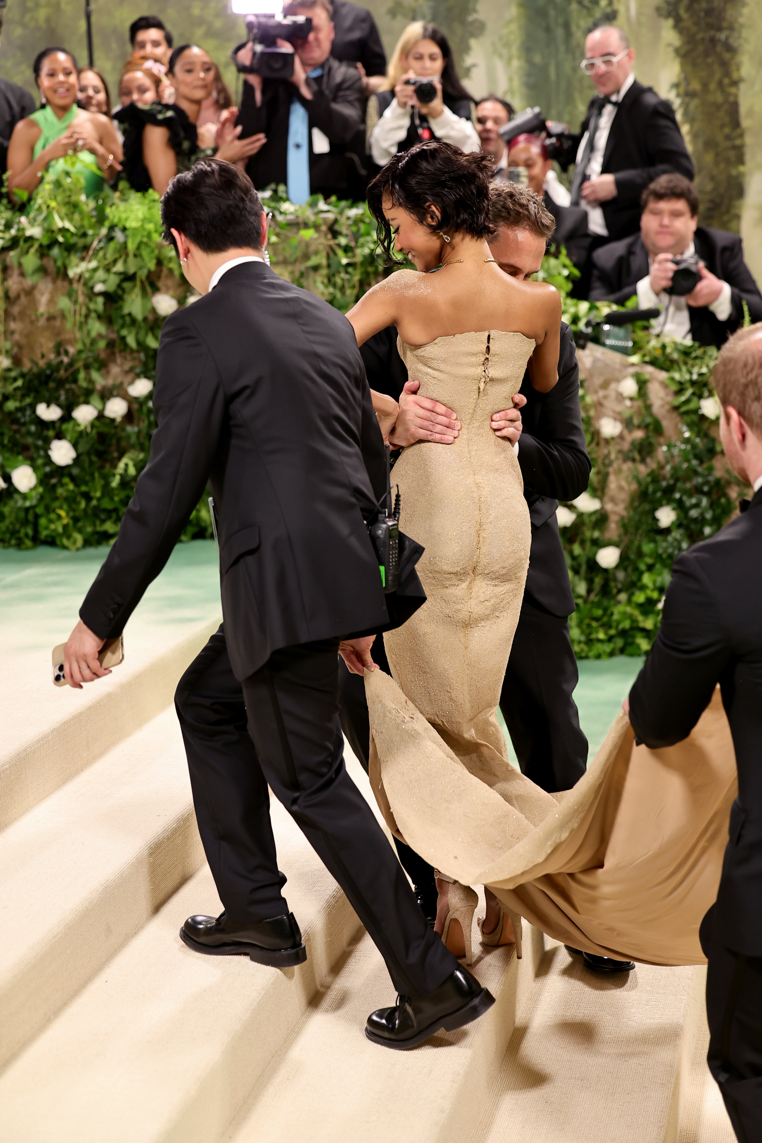 Tyla in an elegant backless gown with a flowing train being lifted by a man as two others carry her train