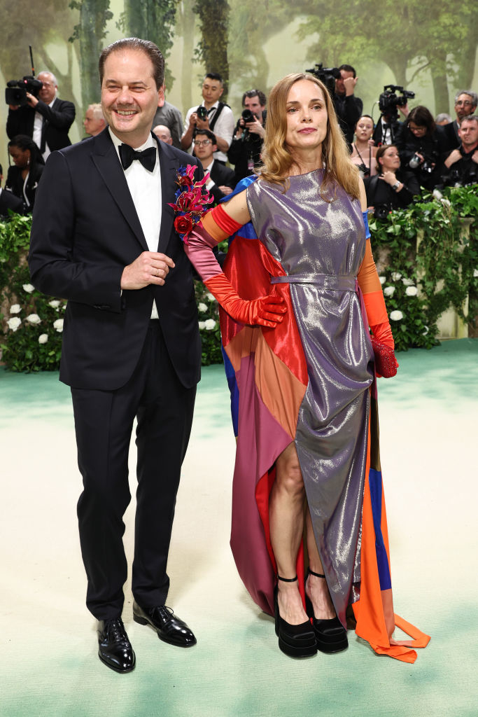 Max wearing a suit with a floral accessory, Nina in a metallic, color-block dress, pose at an event with photographers in the background