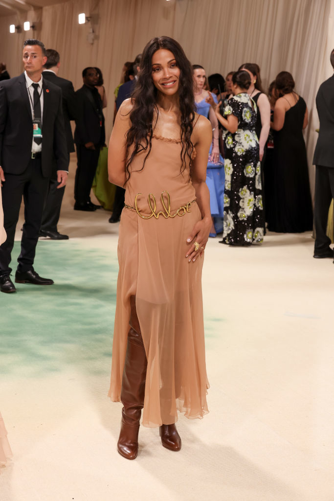 Zoe Saldana in a sheer sleeveless dress with metallic belt detail, posing on a carpeted area. Other attendees in background