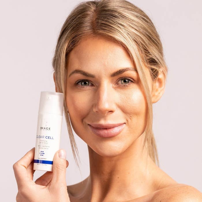 Woman holding skincare product, looking at camera, neutral background, for shopping content